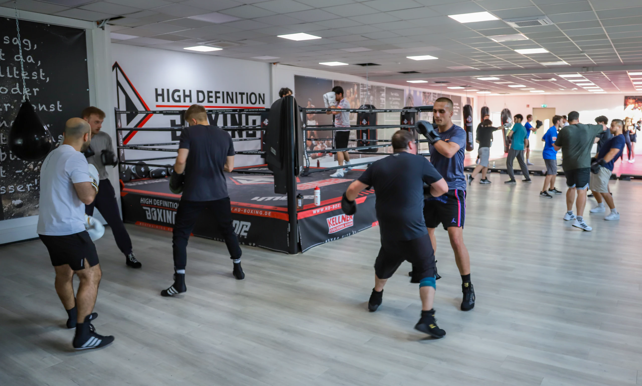 Volle Trainingshalle mit Boxring, Mattenfläche und mehreren Trainingspaaren
